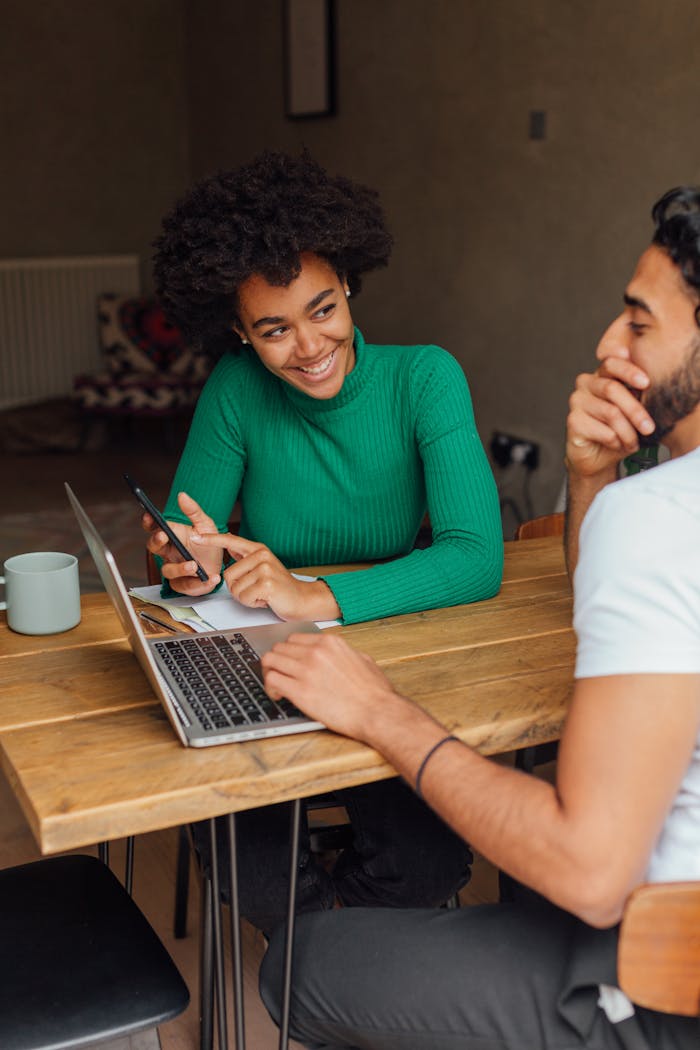 Two adults collaborating at a wooden table with laptops and phones in a cozy indoor setting.